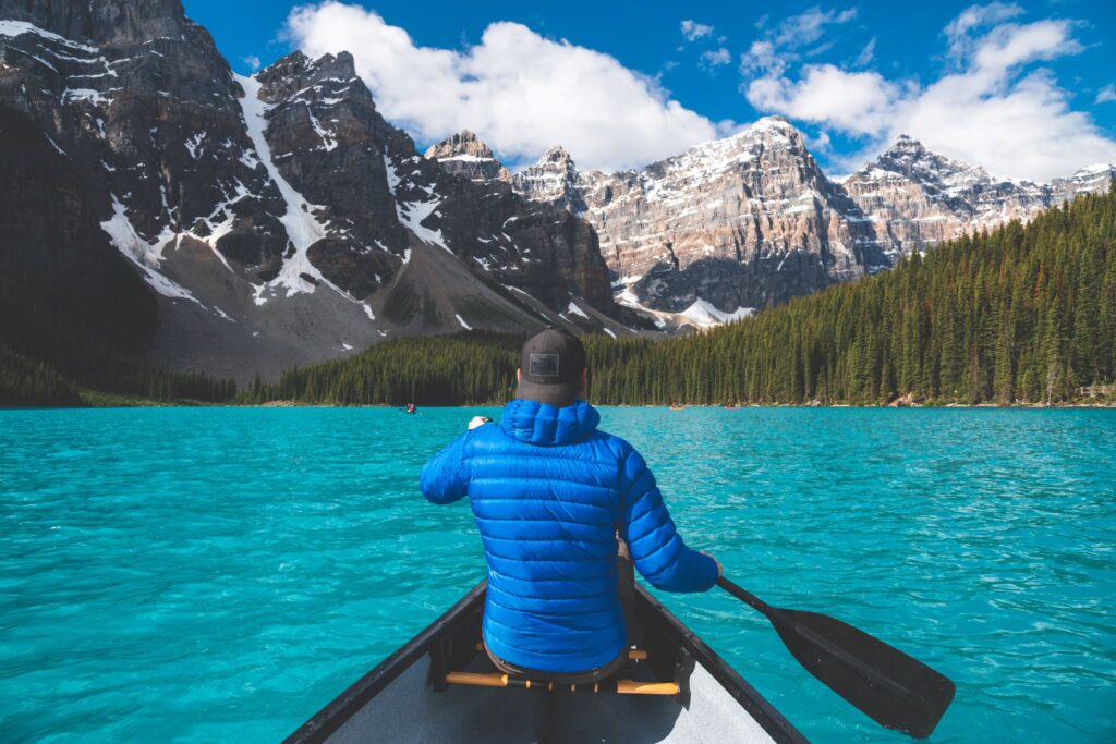 Photo of man from behind canoeing in a blue lake in front of mountains and a tree line in Alberta.