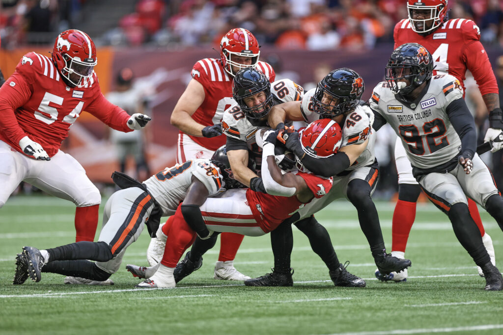 Football players from the BC Lions tackling a player on the Calgary Stampeders team during a game.